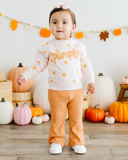 Baby wearing pink crewneck sweatshirt with pumpkin pattern and patch that reads 'pumpkin' and orange pants
