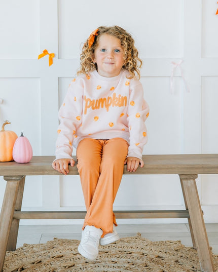 Girl sitting on a bench wearing pink crewneck sweatshirt with pumpkin pattern and patch that reads 'pumpkin' and orange pants