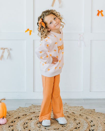 Girl wearing pink crewneck sweatshirt with pumpkin pattern and patch that reads 'pumpkin' and orange pants