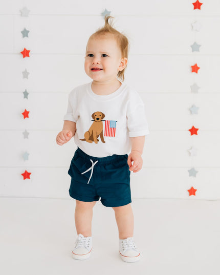 Baby wearing white romper with dog holding American flag and blue shorts