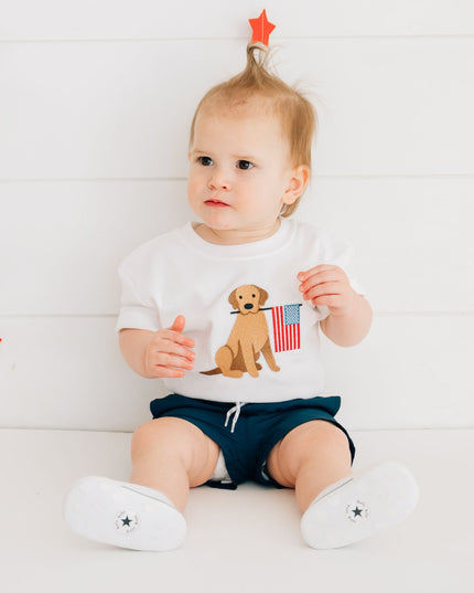 Baby wearing white romper with dog holding American flag and blue shorts