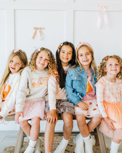 Five young girls sitting on a bench wearing matching outfits with pumpkins.