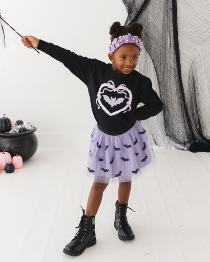 A girl wearing a black sweatshirt with a purple bat, purple tutu with bats and a bat wand. 