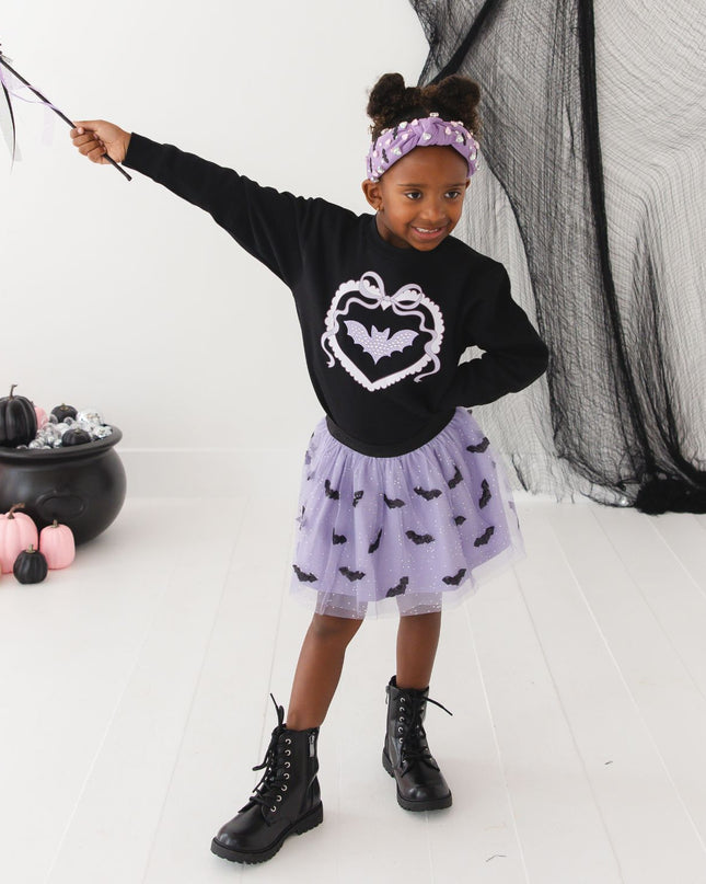 A girl wearing a black sweatshirt with a purple bat, purple tutu with bats and a bat wand. 