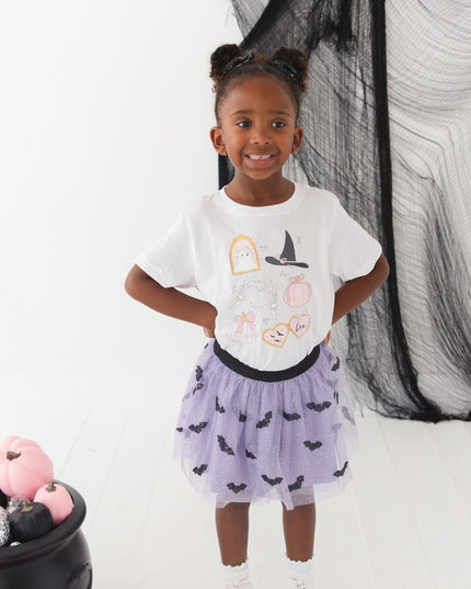 A girl wearing a white short-sleeve t-shirt with cute Halloween-themed graphics and a purple tutu with bats