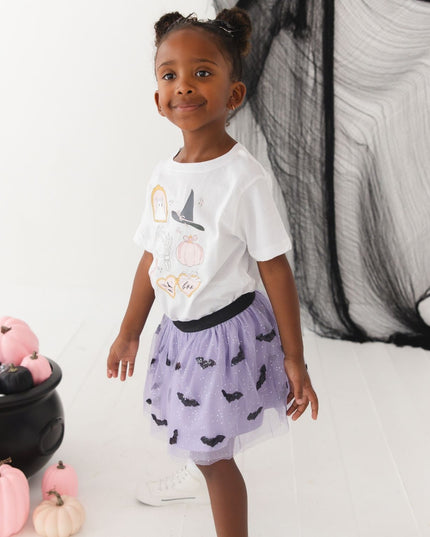 A girl wearing a white short-sleeve t-shirt with cute Halloween-themed graphics and a purple tutu with bats