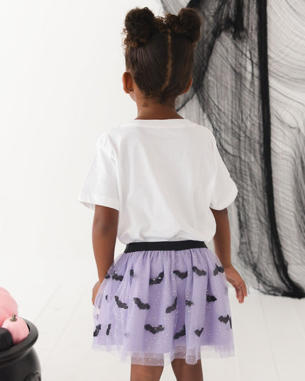 A girl wearing a white short-sleeve t-shirt with cute Halloween-themed graphics and a purple tutu with bats
