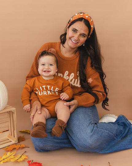 A woman and baby sitting together  wearing matching rust-orange tops - the baby's says 'little turkey' and the woman's says 'thankful
