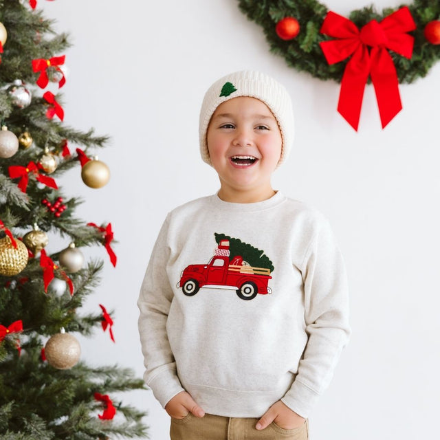 Boy wearing a natural color sweatshirt with red truck carrying a Christmas tree patch and hat with a Christmas tree