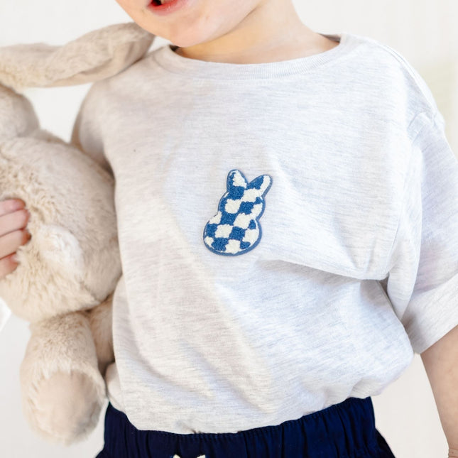 Boy wearing ash grey t-shirt with blue and white bunny checkered patch holding a stuffed bunny