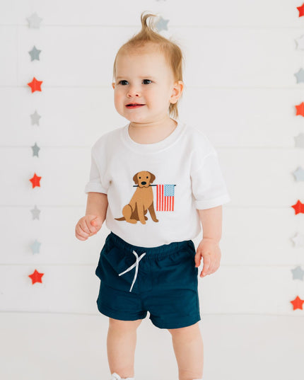 Baby wearing white romper with dog holding American flag and blue shorts