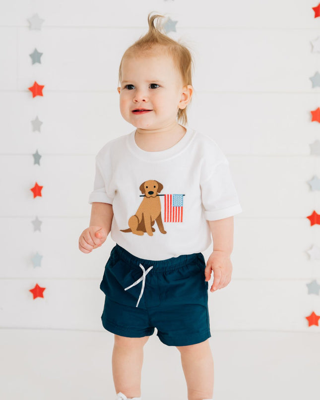 Baby wearing white romper with dog holding American flag and blue shorts