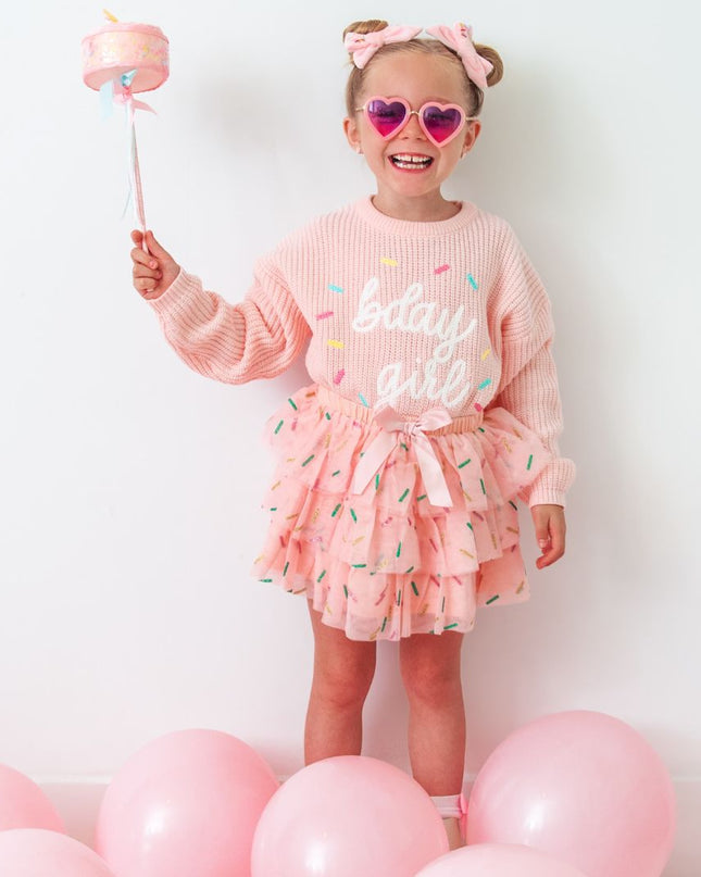 Girl wearing a pink sweater embroidered with 'bday girl' and pink tutu, surrounded by pink balloons
