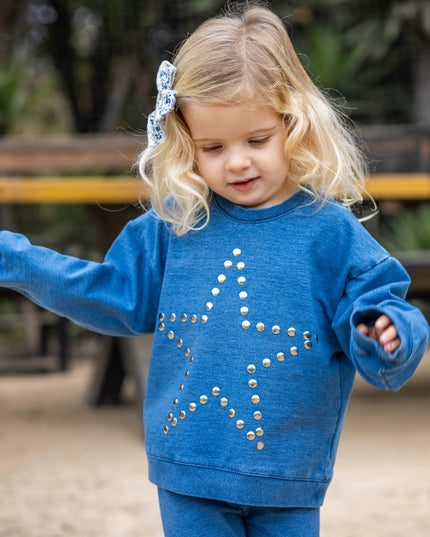 A young blonde girl wearing a matching blue sweatshirt and sweatpants set. The sweatshirt features a large star design outlined in silver studs.