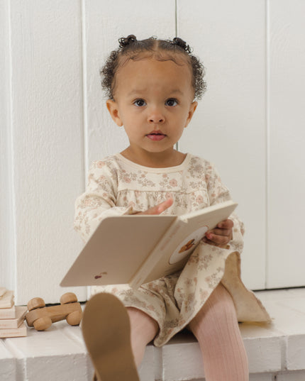 Toddler reading a book wearing a long-sleeve dress with a tiered ruffle hem and floral pattern with pink tights