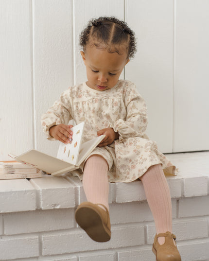 Toddler reading a book wearing a long-sleeve dress with a tiered ruffle hem and floral pattern with pink tights