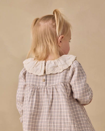 A little girl wearing a long sleeve dress with blue checkered pattern and white collar 