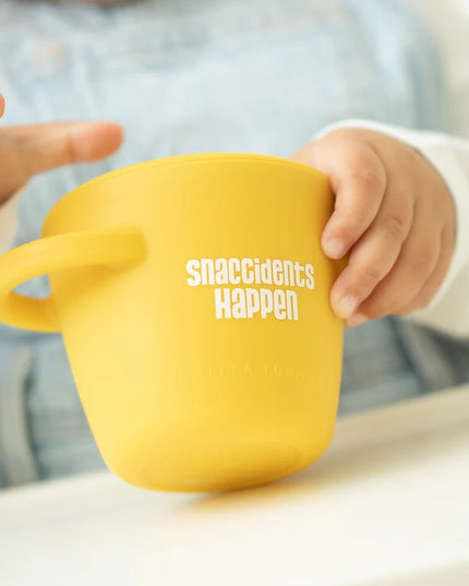 A toddler eating from yellow snack container with lid and handles, featuring the text 'snaccidents Happen' 
