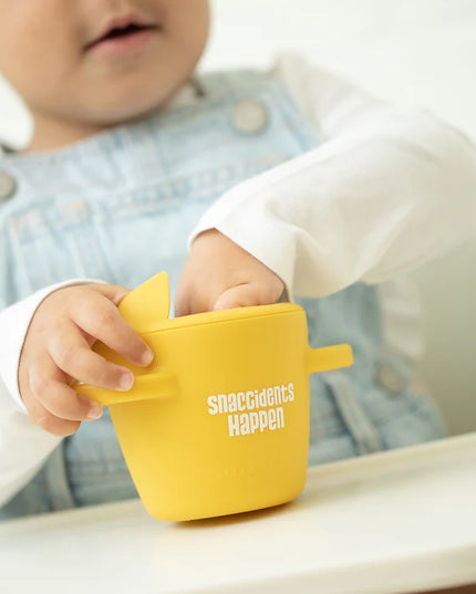 A toddler eating from yellow snack container with lid and handles, featuring the text 'snaccidents Happen' 