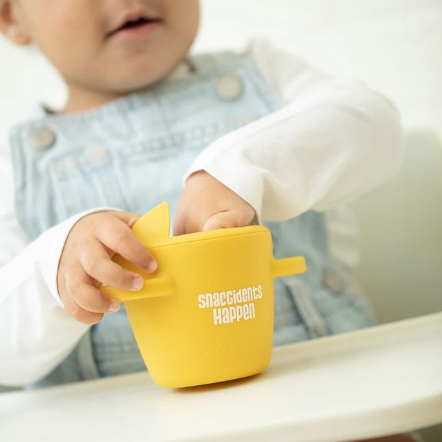 A toddler eating from yellow snack container with lid and handles, featuring the text 'snaccidents Happen' 