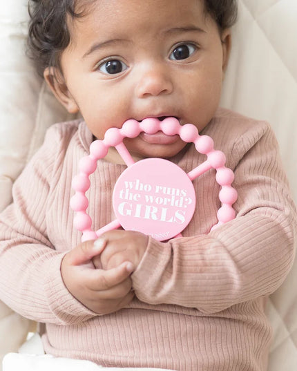 A baby chewing on a pink silicone baby teether with the text 'who runs the world? GIRLS' 