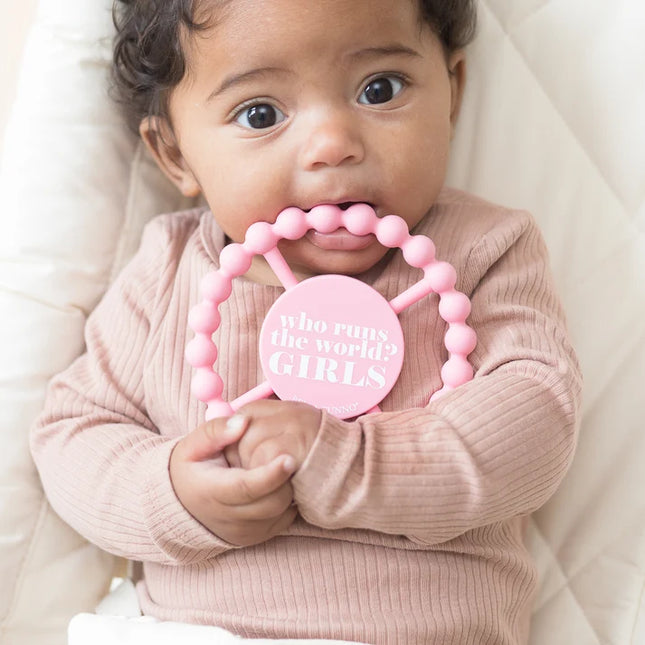 A baby chewing on a pink silicone baby teether with the text 'who runs the world? GIRLS' 