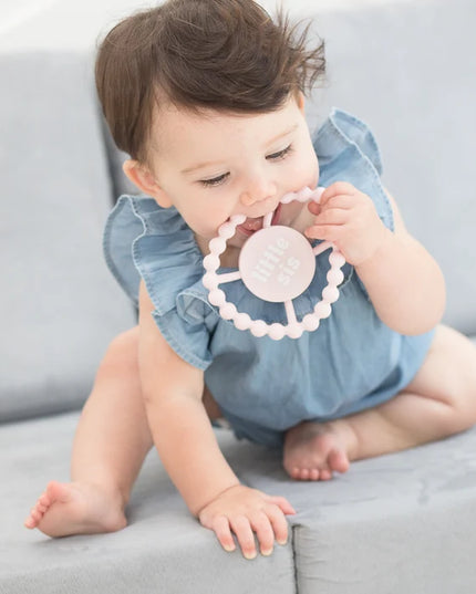 Baby chewing on light pink teething ring with 'little sis' text
