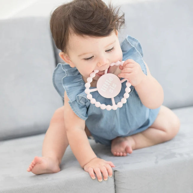 Baby chewing on light pink teething ring with 'little sis' text