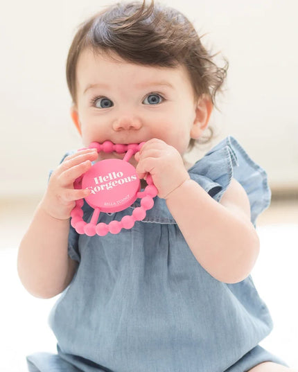 Baby holding pink silicone teething ring with 'hello gorgeous' text