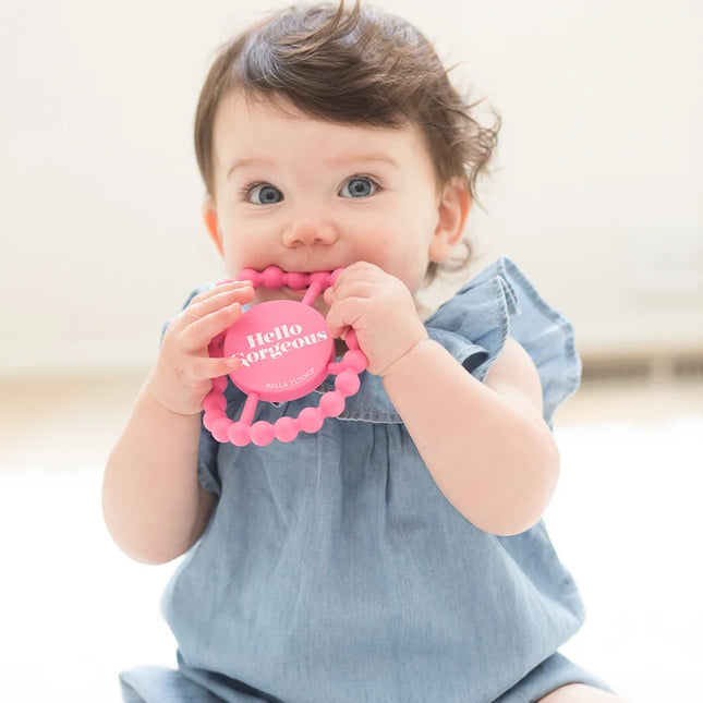 Baby holding pink silicone teething ring with 'hello gorgeous' text