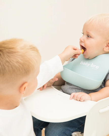 Boy feeding baby wearing a bib with 'baby bro' text