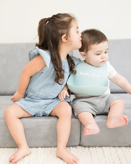 Two children sitting on a couch, one wearing a bib with 'baby bro' text