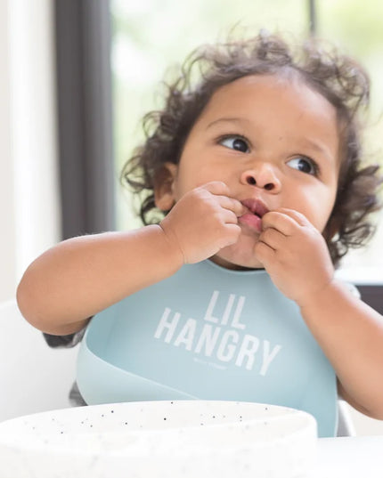 Baby wearing a light blue 'LIL HANGRY' bib while eating.