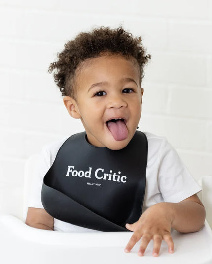 A baby wearing a black silicone baby bib with the words 'Food Critic' printed on the front