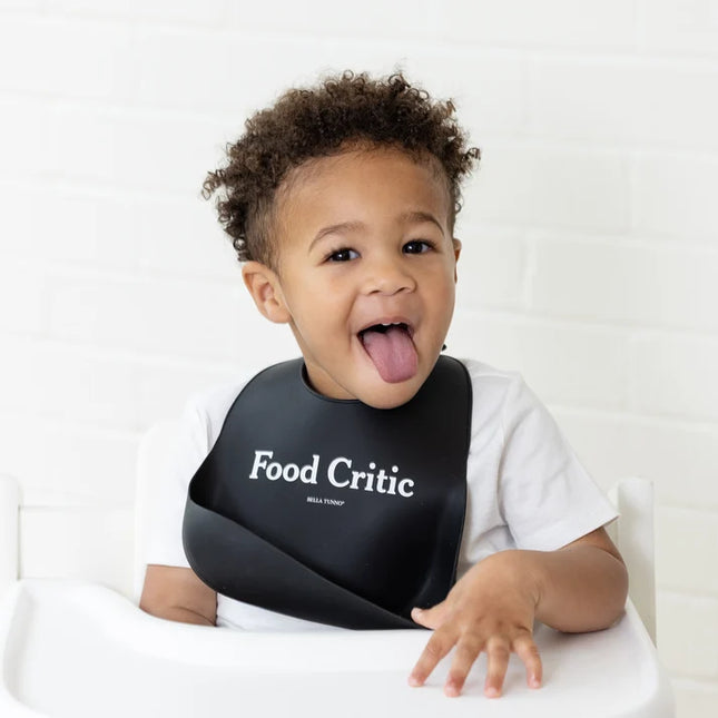 A baby wearing a black silicone baby bib with the words 'Food Critic' printed on the front