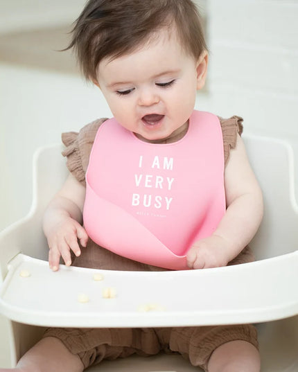 Baby in a high chair wearing a pink bib with 'I AM VERY BUSY' text.