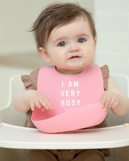 Baby in a high chair wearing a pink bib with 'I AM VERY BUSY' text.
