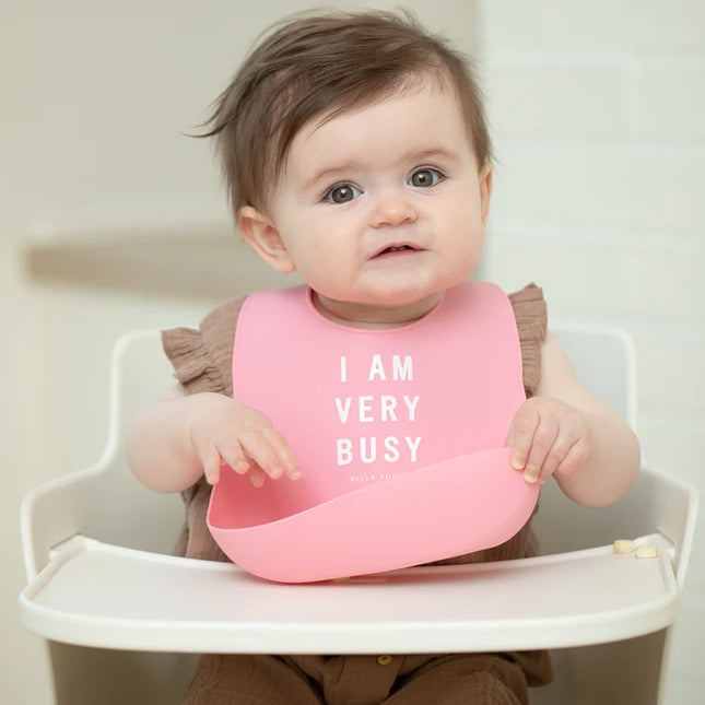 Baby in a high chair wearing a pink bib with 'I AM VERY BUSY' text.
