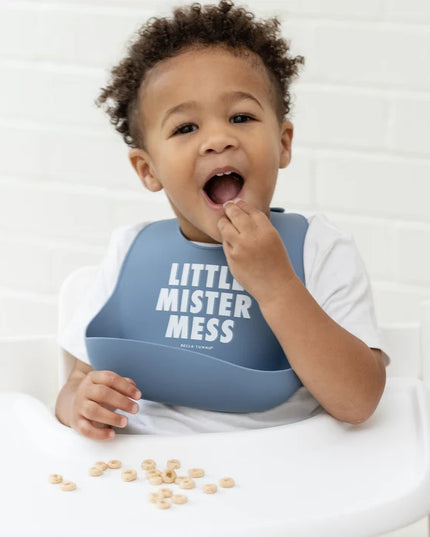 Baby in a high chair eating and wearing blue bib with 'Little Mister Mess' text