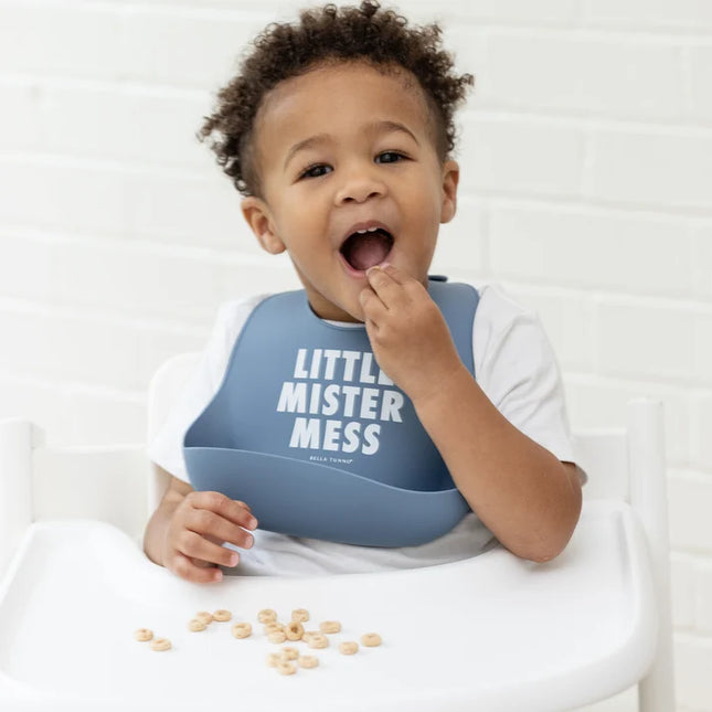 Baby in a high chair eating and wearing blue bib with 'Little Mister Mess' text