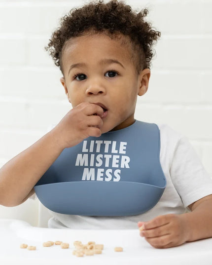 Baby in a high chair eating and wearing blue bib with 'Little Mister Mess' text
