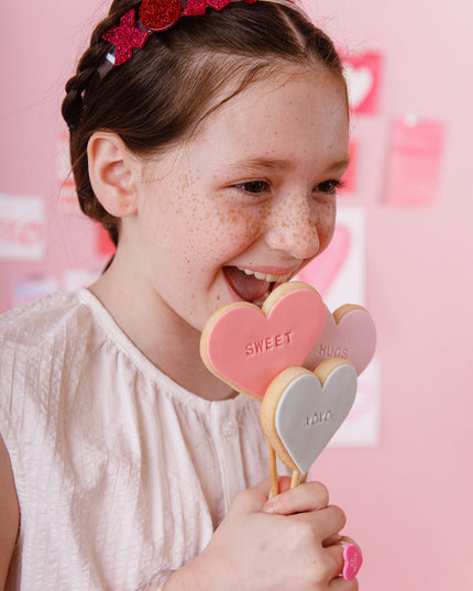 Girl wearing headband with red and pink glitter xoxo design eating cookie lollipops 