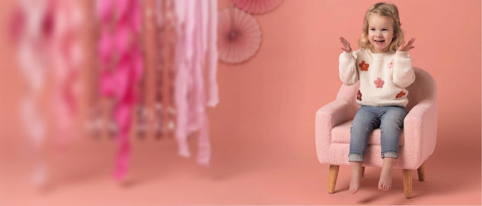 A young girl with blonde hair sits smiling in a pink armchair against a solid pink background. She is wearing a cream ribbed knit sweater decorated with orange and pink floral patches, paired with blue jeans.